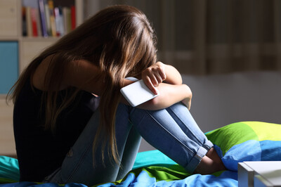 Zu sehen ist ein junges Mädchen / eine junge Frau, die mit dem Handy in der Hand auf ihrem Bett sitzt, den Kopf auf die Arme legt und verzweifelt wirkt.