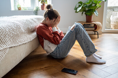 Mädchen sitzt vor ihrem Bett und hat den Kopf zwischen den Händen verborgen. Neben ihr liegt ein Handy.