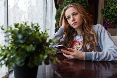 Junges Mädchen mit Smartphone in der Hand schaut traurig aus dem Fenster