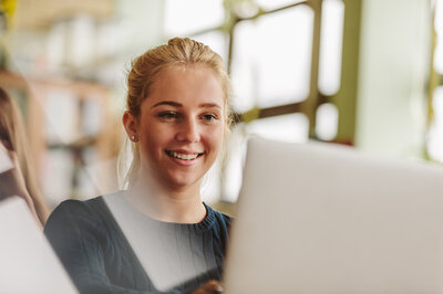 Eine junge Frau sitzt lächelnd vor einem Laptop.