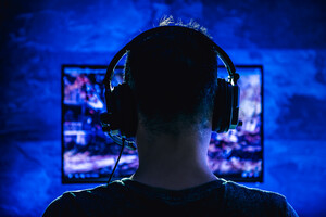 Zu sehen ist eine Person vor einem Gaming-Setup in einem halbdunklen Raum. Die Person ist von hinten zu sehen.