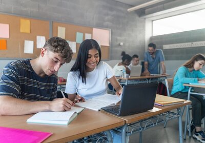 Zwei Schüler:innen sitzen im Klassenzimmer vor einem Laptop und lachen.