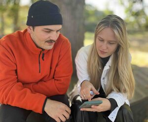 Zu sehen ist eine junge Frau, die einem älteren Mann etwas auf ihrem Handy zeigt. Die beiden sitzen auf einer Parkbank, im Hintergrund ist ein Baum und Wiese zu sehen.