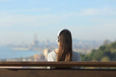 Eine junge Frau ist von hinten zu sehen. Sie sitzt auf einer Bank, trägt Kopfhörer und guckt in die Ferne, wo verschwommen die Skyline einer Stadt zu sehen ist.