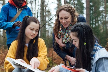 Drei Schüler:innen und eine Lehrer:in im Wald und schauen auf ein Buch und ein Handy.