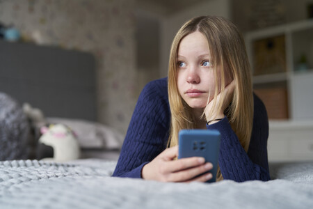 Einsamkeit, Isolation und Verlust von digitalen Angeboten. Was bringt das Social Media Verbot? Eine junge Frau liegt auf dem Bett mit Handy in der Hand und schaut betrübt zur Seite.