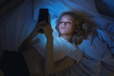 Eine junge Frau liegt im Bett auf dem Rücken und schaut auf ihr Handy. Das Displaylicht beleuchtet das Gesicht der Frau.