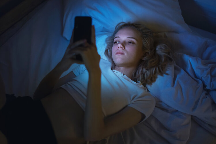Eine Junge Frau liegt im Bett und schaut auf ihr Handy. Sie wird vom Displaylicht angestrahlt.
